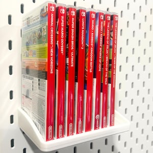 Könnte beinhalten: Ein weißes Regal mit acht roten Nintendo Switch-Spielkassetten. Die Spiele sind: Animal Crossing: New Horizons, Super Mario Odyssey, Super Smash Bros. Ultimate, Luigi's Mansion 3, Mario + Rabbids: Kingdom Battle, Mario + Rabbids: Sparks of Hope, The Legend of Zelda: Breath of the Wild und The Legend of Zelda: Tears of the Kingdom.