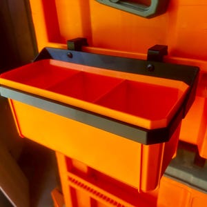 May include: An orange plastic tool organizer with a black trim and three compartments. The organizer is attached to a larger orange structure with black brackets. The image is well-lit, highlighting the product's design and functionality.
