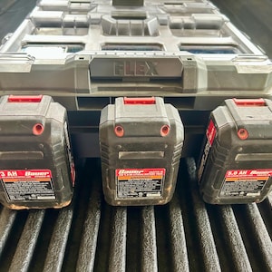 May include: A gray plastic FLEX storage case with a transparent lid positioned behind three black Bauer lithium-ion battery packs. Each battery pack features red accents, a warning label, and specific capacity markings of 3 AH and 5.0 AH.