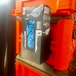 May include: A black box of 100 medium-duty disposable gloves with blue gloves visible through a cutout. The box has the word "GLOVES" and a hand holding a wrench printed on it. The box is next to orange toolboxes.