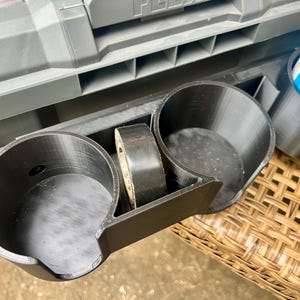 Peut inclure: Un porte-gobelet en plastique noir imprim&eacute; en 3D dot&eacute; de deux compartiments circulaires et d'une fente centrale pour un rouleau de ruban adh&eacute;sif noir. L'accessoire est fix&eacute; &agrave; une bo&icirc;te &agrave; outils grise, avec une bombe de liquide de d&eacute;marrage bleue.
