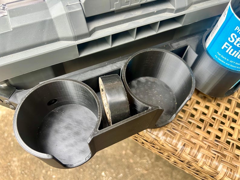 Peut inclure: Un porte-gobelet en plastique noir imprim&eacute; en 3D dot&eacute; de deux compartiments circulaires et d'une fente centrale pour un rouleau de ruban adh&eacute;sif noir. L'accessoire est fix&eacute; &agrave; une bo&icirc;te &agrave; outils grise, avec une bombe de liquide de d&eacute;marrage bleue.