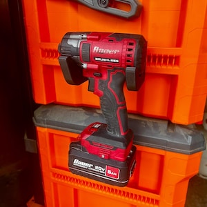 May include: A red and black Bauer brushless impact wrench with a 20V 6Ah battery. The tool is set against a backdrop of orange storage bins. The tool's handle has a textured grip for secure handling.