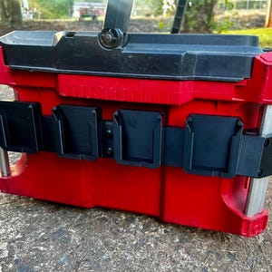 May include: A red and black tool box with a black handle and a black belt with four black holders. The tool box is sitting on a concrete surface. The tool box is designed to hold tools.