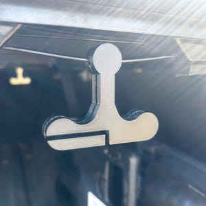 May include: A 3D printed plastic hook designed to hang items from a car's trunk lid. The hook is gray and has a unique shape with a rounded top and two arms extending out.