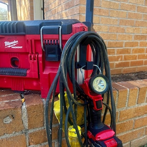 Könnte beinhalten: Eine rote und schwarze Milwaukee Packout Werkzeugkiste mit schwarzem Griff und Verschlüssen. Eine rot-schwarze Arbeitsleuchte mit heller LED ist an einem schwarzen Ständer befestigt, zusammen mit einem aufgerollten schwarzen Kabel. Die Gegenstände stehen auf einer Backsteinmauer.