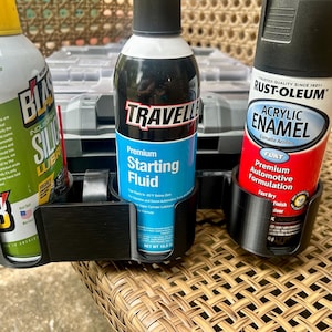 Peut inclure: Un support noir en plastique pour trois bombes a&eacute;rosols contenant : un lubrifiant au silicone Blaster vert, un liquide de d&eacute;marrage Traveller bleu et une peinture &eacute;mail acrylique Rust-Oleum rouge. Le support est pos&eacute; sur une surface tress&eacute;e.