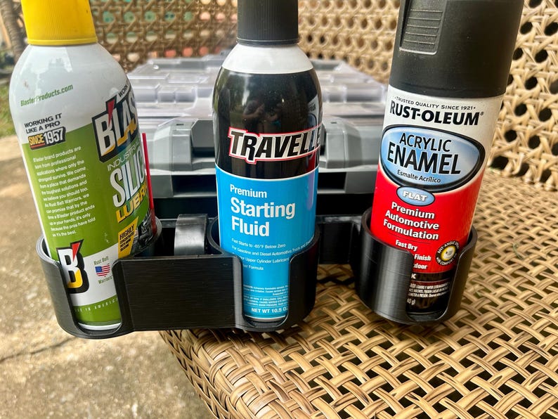 Peut inclure: Un support noir en plastique pour trois bombes a&eacute;rosols contenant : un lubrifiant au silicone Blaster vert, un liquide de d&eacute;marrage Traveller bleu et une peinture &eacute;mail acrylique Rust-Oleum rouge. Le support est pos&eacute; sur une surface tress&eacute;e.