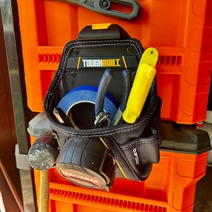 May include: Black and yellow ToughBuilt tool belt with various tools, including pliers, tape measure, and a yellow utility knife. The tool belt is hanging on an orange storage container.