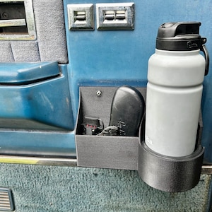Puede incluir: Un organizador de almacenamiento para puerta de plástico negro montado en un panel de puerta de vehículo azul. El organizador cuenta con un portavasos cilíndrico que contiene una botella de agua Bubba gris claro y un compartimento.