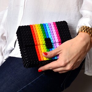 May include: Black clutch with a rainbow stripe design. The clutch is made of small, textured blocks, with a colorful stripe of red, orange, yellow, green, blue, and purple. The clutch is held by a person wearing a white shirt and blue jeans.