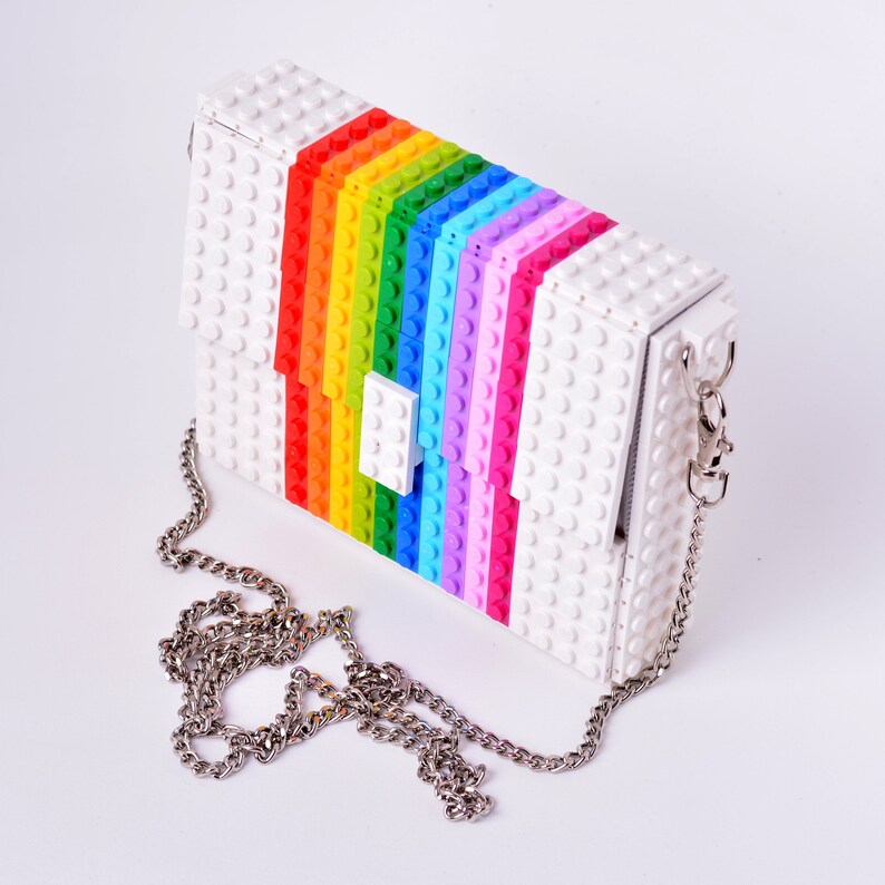 Op de afbeelding: Een witte tas gemaakt van plastic bouwstenen met een regenboogpatroon en een zilveren kettingband.
