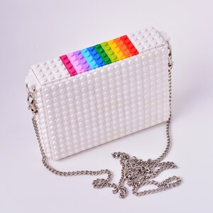 Op de afbeelding: Een witte tas gemaakt van Lego-stenen met een regenboog van gekleurde stenen op de bovenklep. De tas heeft een zilveren kettingband.