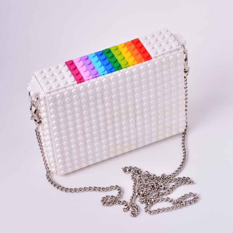 Op de afbeelding: Een witte tas gemaakt van Lego-stenen met een regenboog van gekleurde stenen op de bovenklep. De tas heeft een zilveren kettingband.