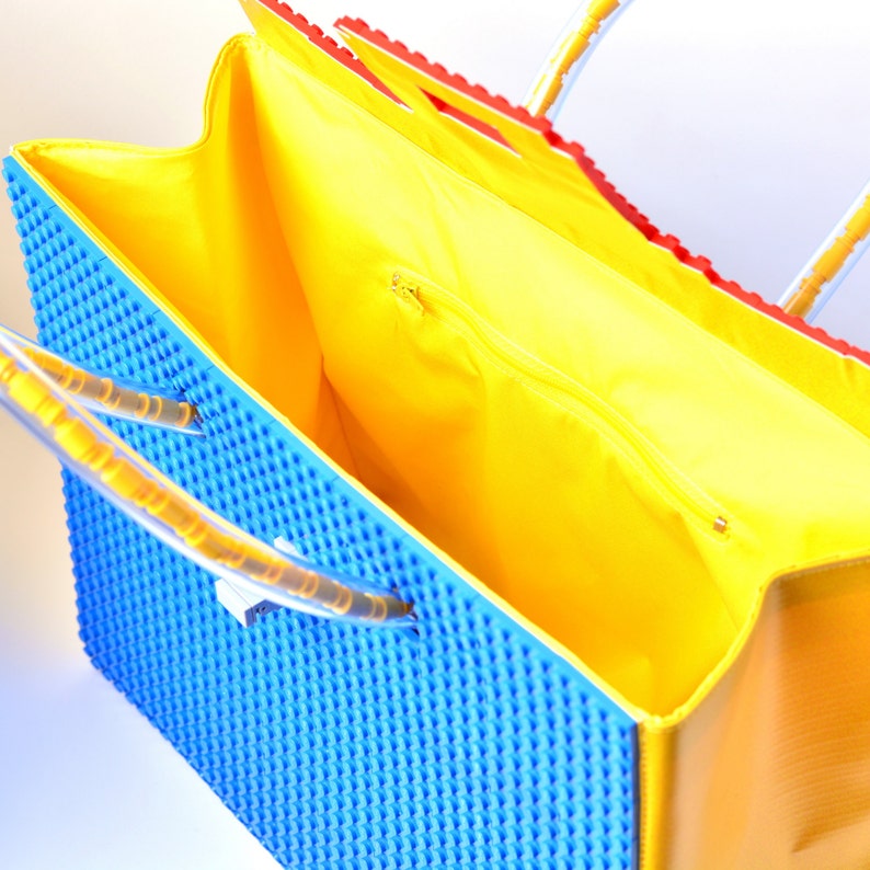 May include: A blue and yellow tote bag with a zippered top and clear plastic handles. The bag is made to look like a Lego brick.