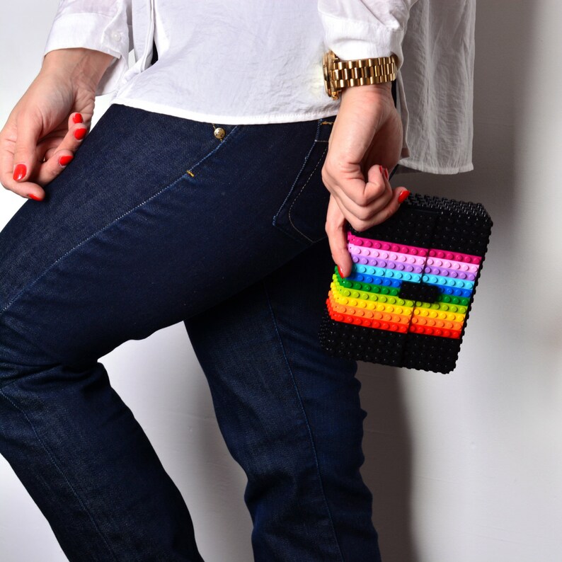 May include: A black clutch made of small, interlocking blocks, with a rainbow stripe design across the front. The clutch is held by a person wearing a white shirt and dark blue jeans.