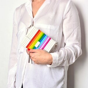 Op de afbeelding: Een witte tas gemaakt van kleurrijke Lego-stenen. De tas heeft een regenboogpatroon en een zilveren kettingband.