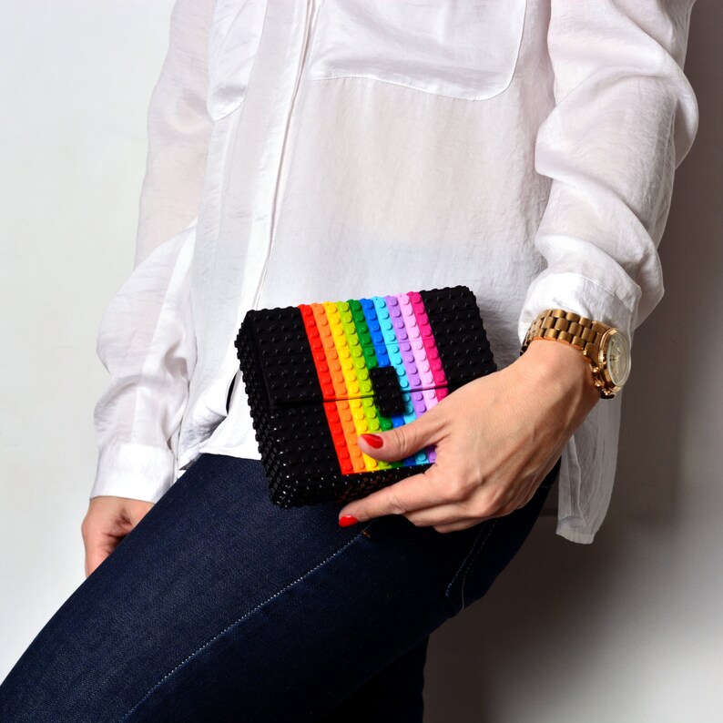 May include: Black clutch with a rainbow stripe design. The clutch is made of small, textured blocks, with a colorful stripe of red, orange, yellow, green, blue, and purple. The clutch is held by a person wearing a white shirt and blue jeans.