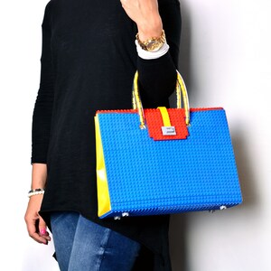 May include: A blue, yellow, and red handbag with a Lego-like design. The bag has a top handle and a flap closure.