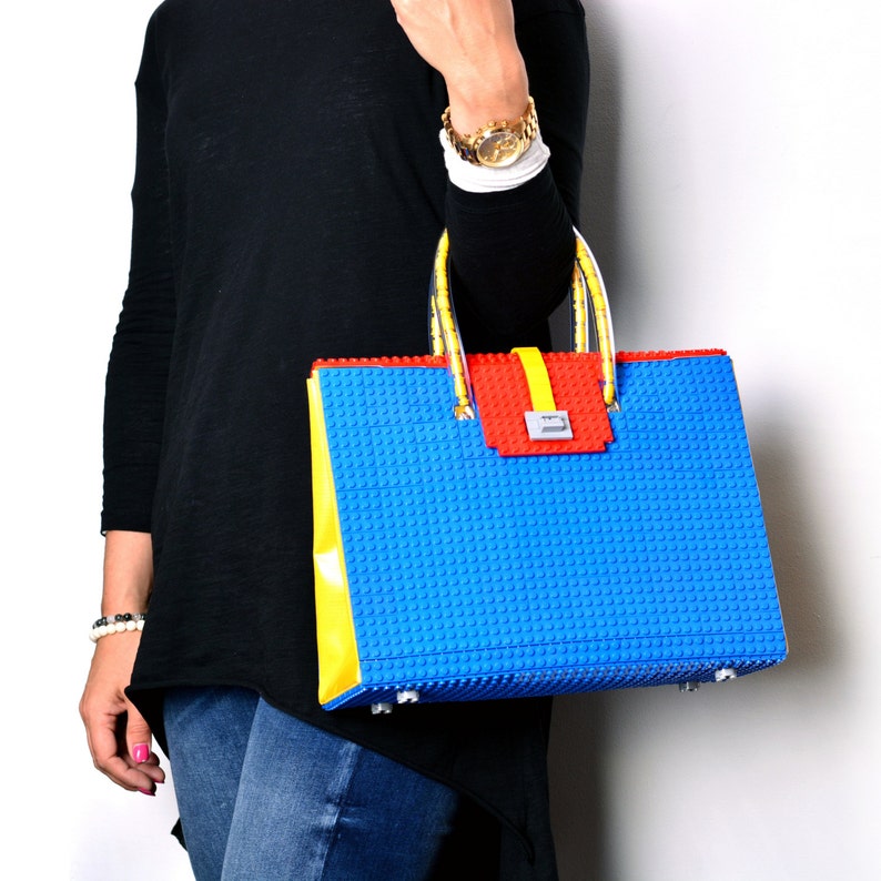 May include: A blue, yellow, and red handbag with a Lego-like design. The bag has a top handle and a flap closure.