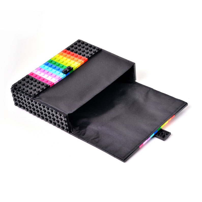 May include: Black clutch with a rainbow-colored brick design on the top flap. The clutch is open, revealing a black interior. The top of the clutch features a rainbow stripe of colorful bricks. The clutch is made of a textured material.