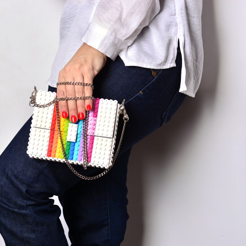 Op de afbeelding: Een witte tas met een regenboogontwerp gemaakt van plastic bouwstenen. De tas heeft een zilveren kettingband.
