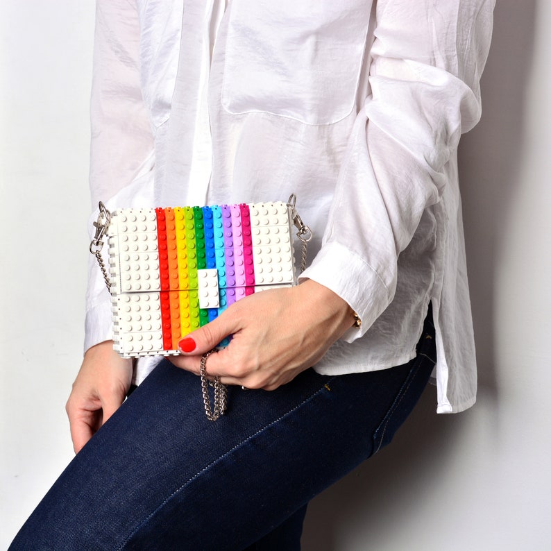 Op de afbeelding: Een witte schoudertas gemaakt van plastic bouwstenen. De tas heeft een regenboogstreep ontwerp en een zilveren kettingriem.