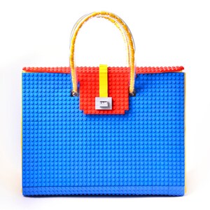 May include: A blue, red, and yellow handbag made of plastic building blocks. The bag has a yellow handle and a red and yellow clasp.