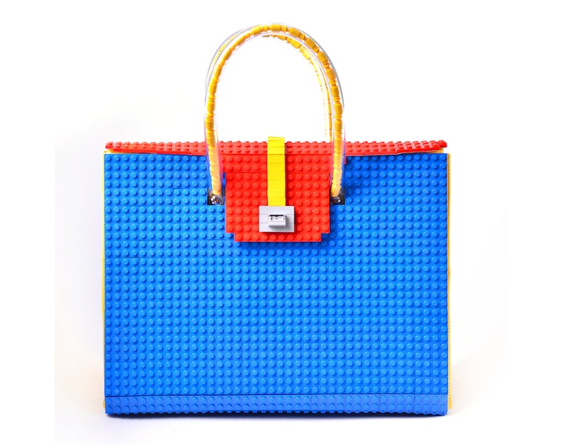 May include: A blue, red, and yellow handbag made of plastic building blocks. The bag has a yellow handle and a red and yellow clasp.