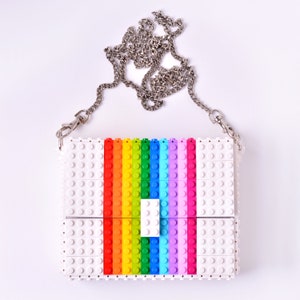 Op de afbeelding: Een witte tas gemaakt van Lego-stenen met een regenboogpatroon. De tas heeft een zilveren kettingband.