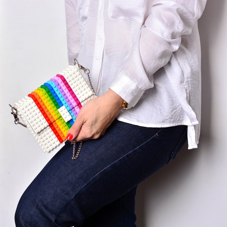 Op de afbeelding: Een witte schoudertas met een regenboogpatroon gemaakt van plastic blokken. De tas heeft een zilveren kettingband en een witte klep met een zilveren gesp.
