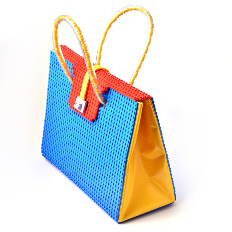 May include: A blue and yellow handbag made of plastic building blocks. The bag has a red flap with a silver clasp and clear plastic handles.
