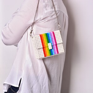 Op de afbeelding: Een witte schoudertas gemaakt van plastic bouwstenen. De tas heeft een regenboogstreep ontwerp en een zilveren kettingband.