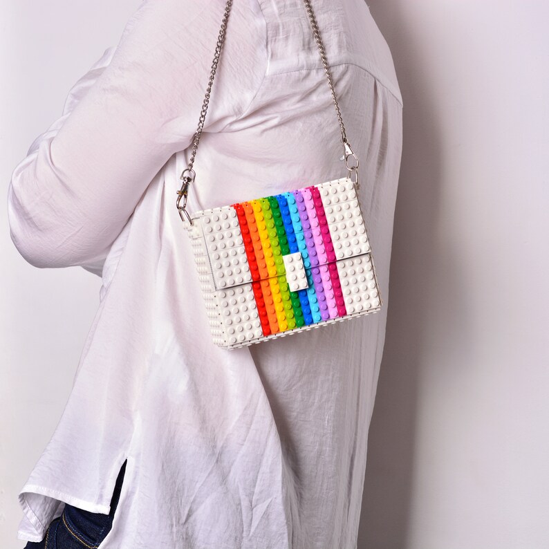 Op de afbeelding: Een witte schoudertas gemaakt van plastic bouwstenen. De tas heeft een regenboogstreep ontwerp en een zilveren kettingband.