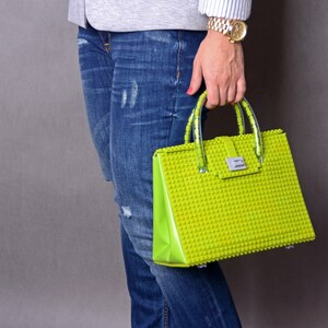 Peut inclure: Un sac &agrave; main vert vif compos&eacute; de pi&egrave;ces en plastique qui s'embo&icirc;tent, ressemblant &agrave; des briques Lego. Le sac a une poign&eacute;e sup&eacute;rieure et un fermoir argent&eacute;.
