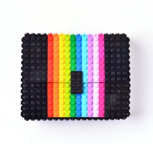 May include: A rectangular clutch made of black and colorful toy building blocks. The center features a rainbow gradient of red, orange, yellow, green, blue, and pink blocks. The clutch is a unique and playful accessory.