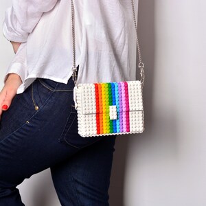 Op de afbeelding: Een witte schoudertas met een regenboogstreepontwerp. De tas heeft een zilveren kettingband en een witte sluiting.