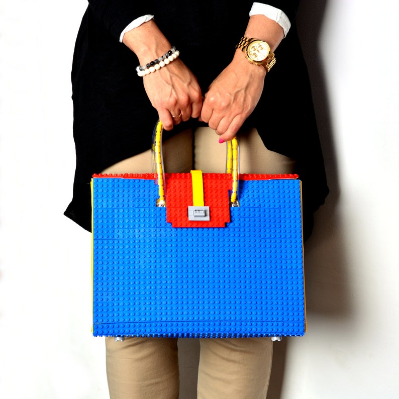 May include: A blue, yellow, and red tote bag made of plastic building blocks. The bag has a top handle and a clasp closure.