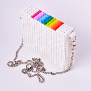 Op de afbeelding: Een witte clutch tas gemaakt van Lego-stenen met een regenboogstreep van Lego-stenen aan de bovenkant. De tas heeft een zilveren kettingband.