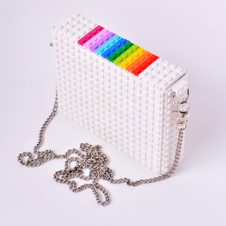 Op de afbeelding: Een witte clutch tas gemaakt van Lego-stenen met een regenboogstreep van Lego-stenen aan de bovenkant. De tas heeft een zilveren kettingband.