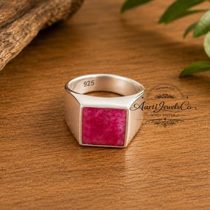 Op de afbeelding: Een zilveren ring met een vierkante, dieproze steen. De ring is gestempeld met "925" en heeft de tekst "HANDMADE Aarti Jewels Co 925 SILVER". De ring ligt op een houten oppervlak.