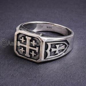 May include: A silver ring with a Jerusalem cross design. The ring features a square face with a large cross and four smaller crosses. The band has a shield-shaped design with a cross. The ring is marked with "925".