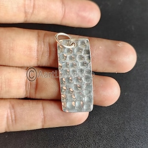 Puede incluir: Un colgante rectangular de plata con textura martillada. El colgante tiene un pequeño lazo en la parte superior para una cadena. La superficie del colgante está cubierta con un patrón de pequeñas hendiduras irregulares.
