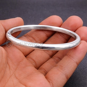 May include: A polished silver bangle bracelet with a smooth, rounded design. The jewellery is held in a hand against a dark background. The bracelet is a simple, elegant piece.