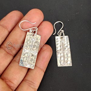 May include: A pair of silver-colored rectangular earrings with a hammered texture. Each earring features a cross design with small decorative elements. The earrings have a hook closure.