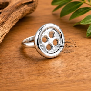 May include: A polished silver ring featuring a circular button-shaped centerpiece with four holes. The ring is engraved with the text Aarti JewelsCo 925 SILVER. The design is smooth and metallic, reflecting light across its rounded surface.