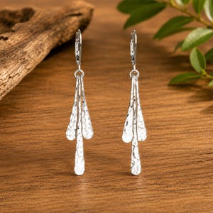 May include: A pair of silver-toned dangle earrings featuring three elongated, hammered metal drops suspended from a leverback closure. The textured surface of the metal reflects light, creating a shimmering effect on each of the three tiered, teardrop-shaped components that hang vertically from the earring wire.