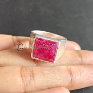 Könnte beinhalten: Ein silberner Ring mit einem großen, quadratischen, rubinroten Edelstein. Der Ring hat ein schlichtes, modernes Design mit einem dicken Band und einer Lünettenfassung. Der Ring wird auf einer Hand präsentiert.