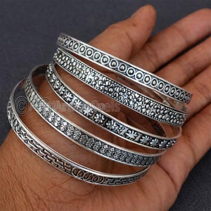 Puede incluir: Un conjunto de pulseras de plata con intrincados diseños, incluyendo círculos, puntos y motivos florales. Las pulseras se muestran en una mano, mostrando su detallada artesanía y diversas texturas. La joyería es probablemente de plata de ley.