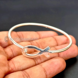 Puede incluir: Una pulsera de brazalete plateada con un símbolo de pez estilizado. La pulsera tiene un diseño simple y circular con el emblema del pez en el centro. La joya se muestra sobre un fondo oscuro.